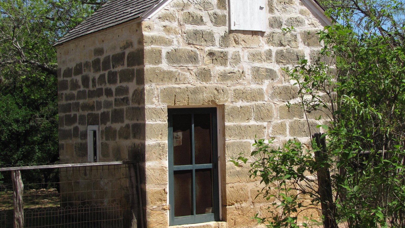 A tall, limestone building with a shingled roof has a screen door entry.
