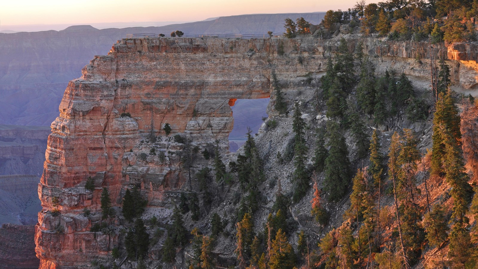 Pink rays of sun illuminate the side of a large, cream-colored stone arch on the side of a cliff.