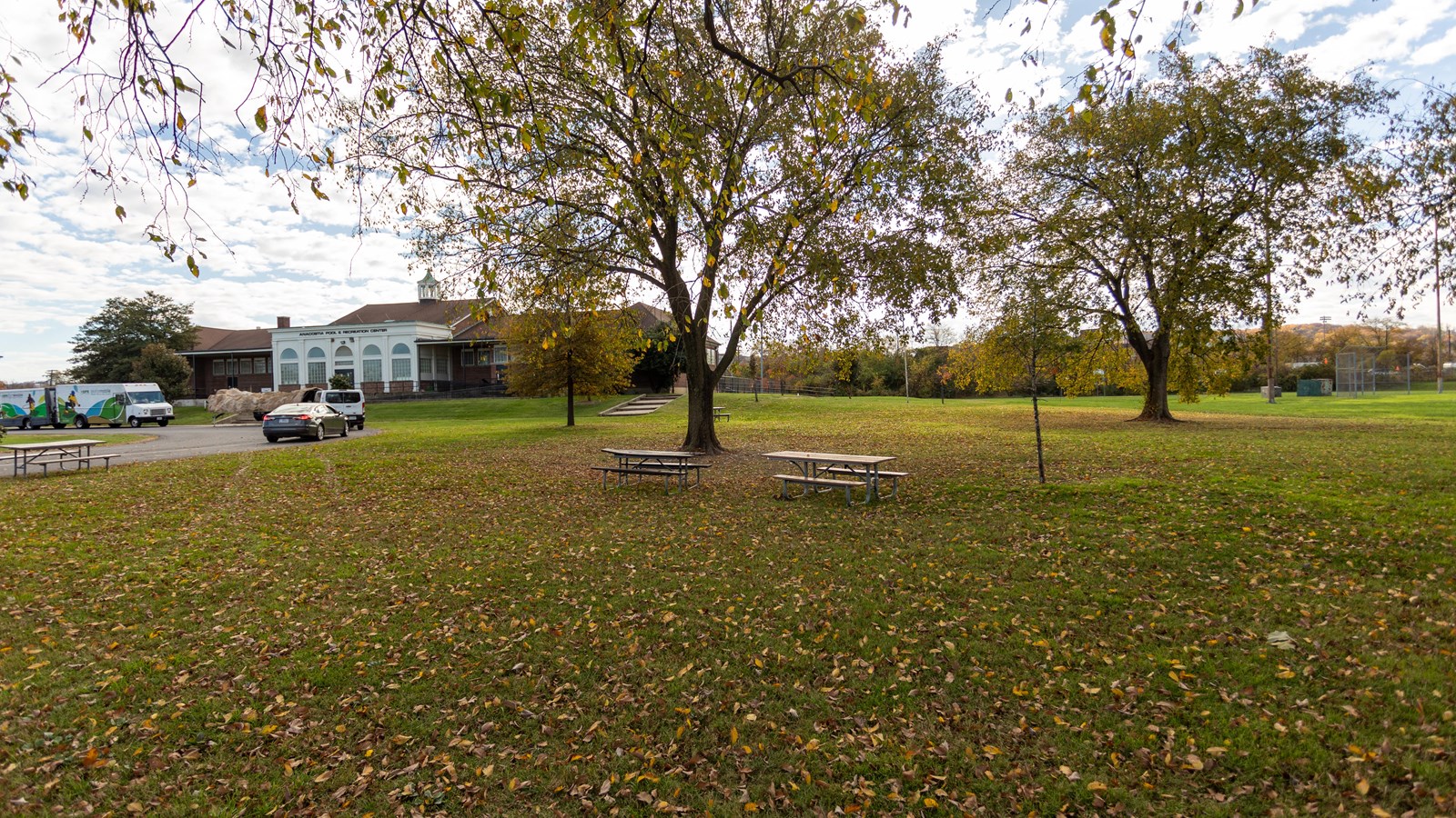 Two picnic tables sit in an open field. A white building with a brick roof is in the background.