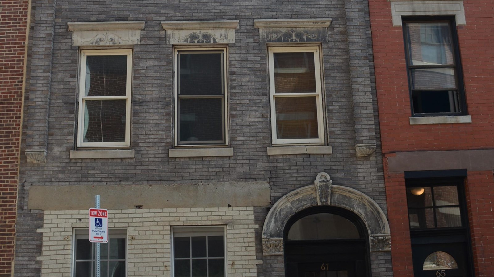 A multi-story grey building with three windows on each floor and a brick building on either side.