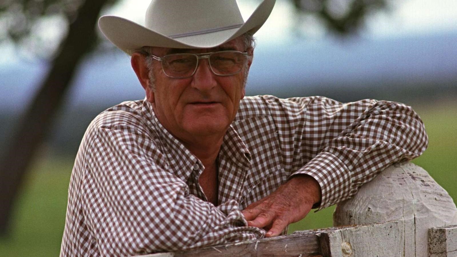 Bespectacled in a cowboy hat and checkered shirt, the president leans on a white fence post.