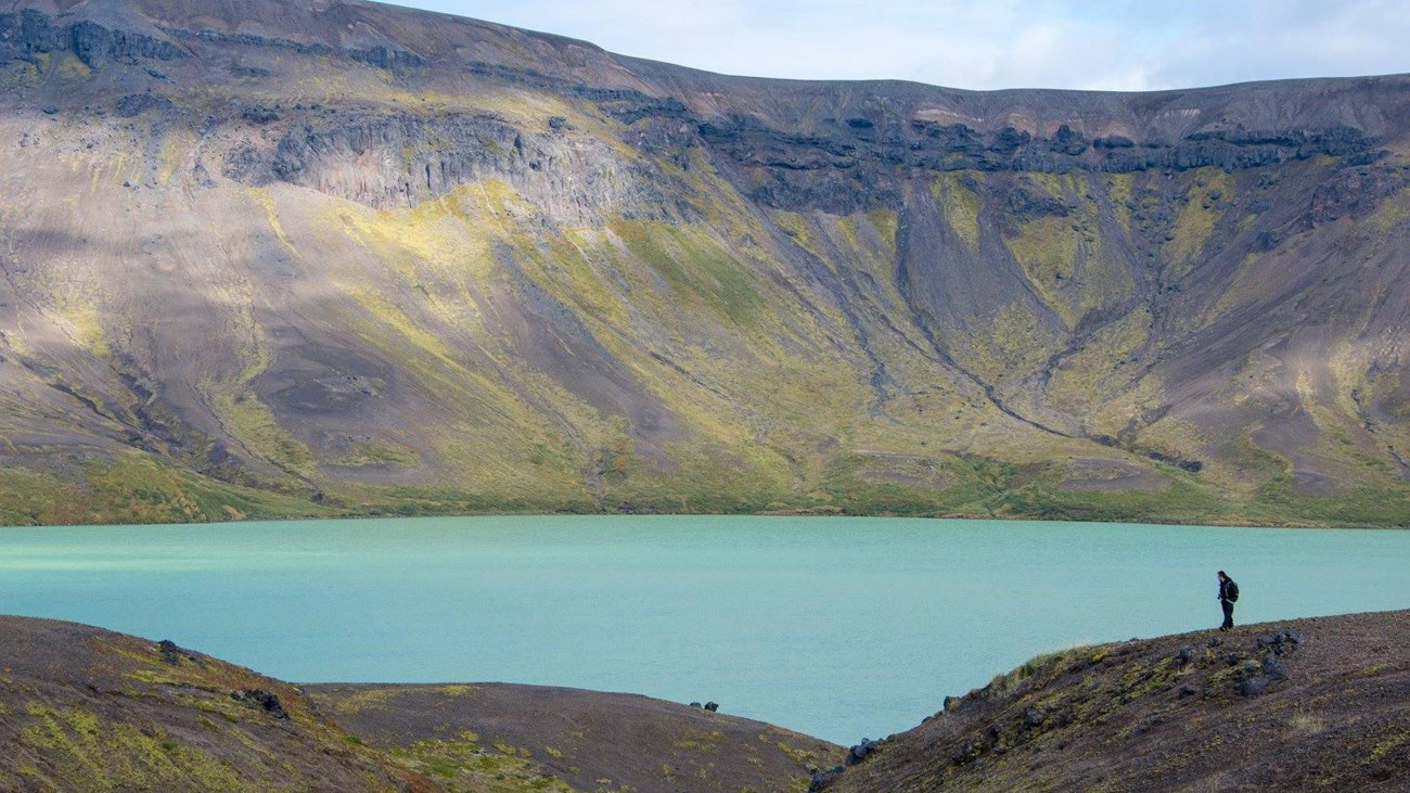 A silhouette of a figure in the distance with a blue lake and caldera rim behind them.
