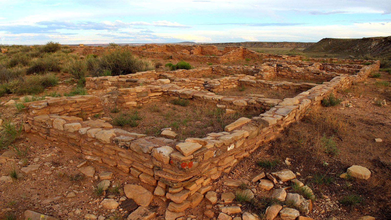 Puerco Pueblo masonry walls just after sunset with a partly cloudy sky, mesa in the background.