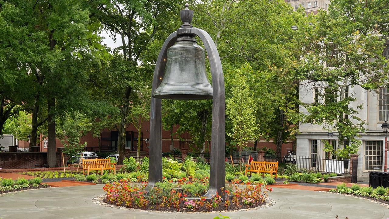 Large bell suspended by an arch surrounded by footpaths and foliage.