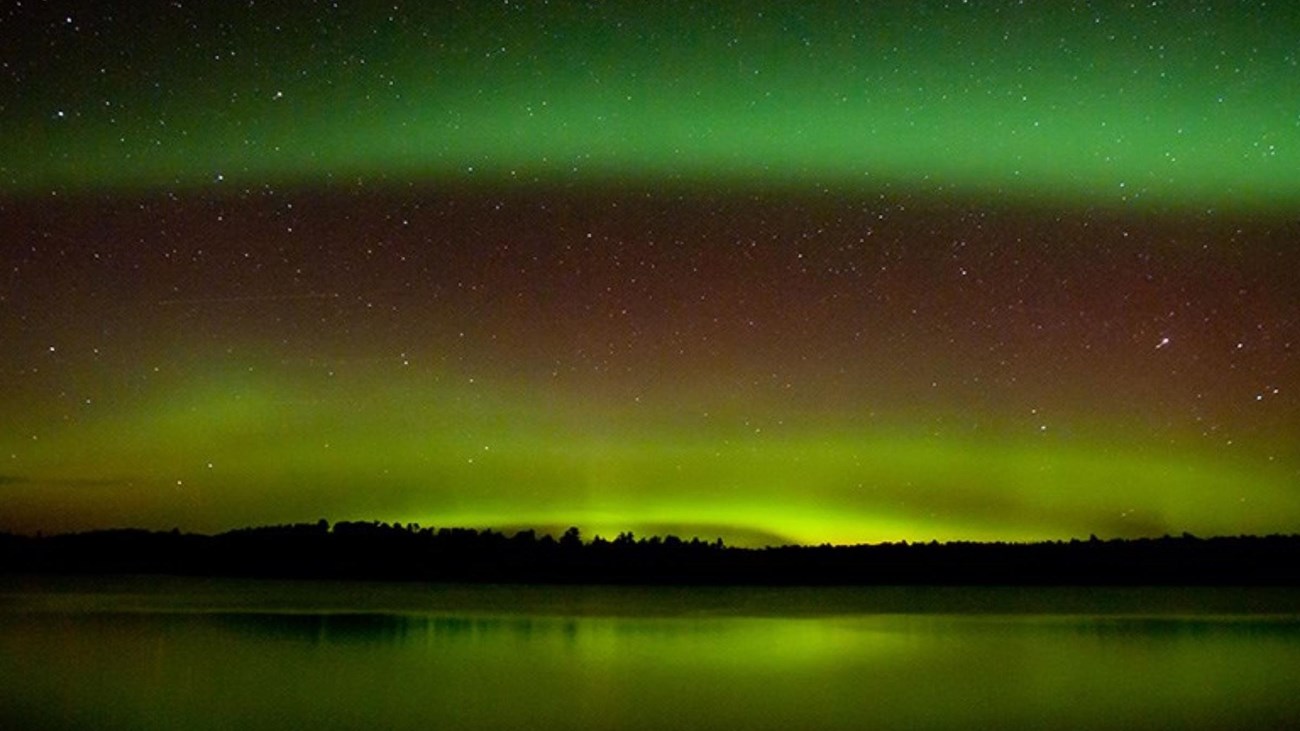 Green waves of light shine in two waves in a dark sky, illuminated the tree line and lake below.