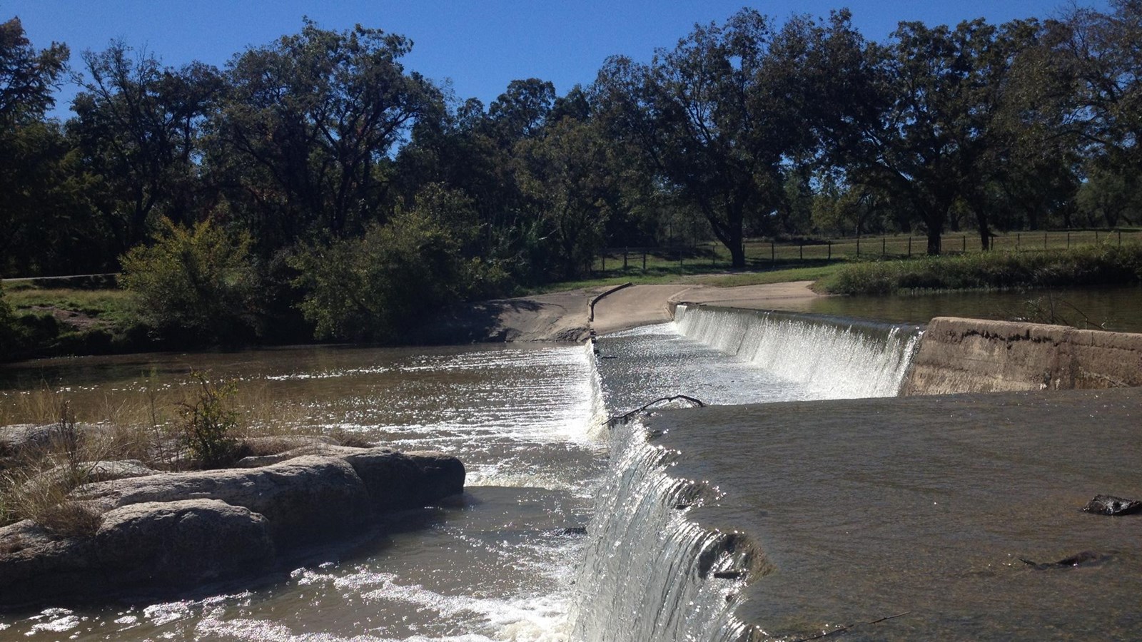 With full trees on the opposite bank, river water flows across a wide, low road creating waterfalls.