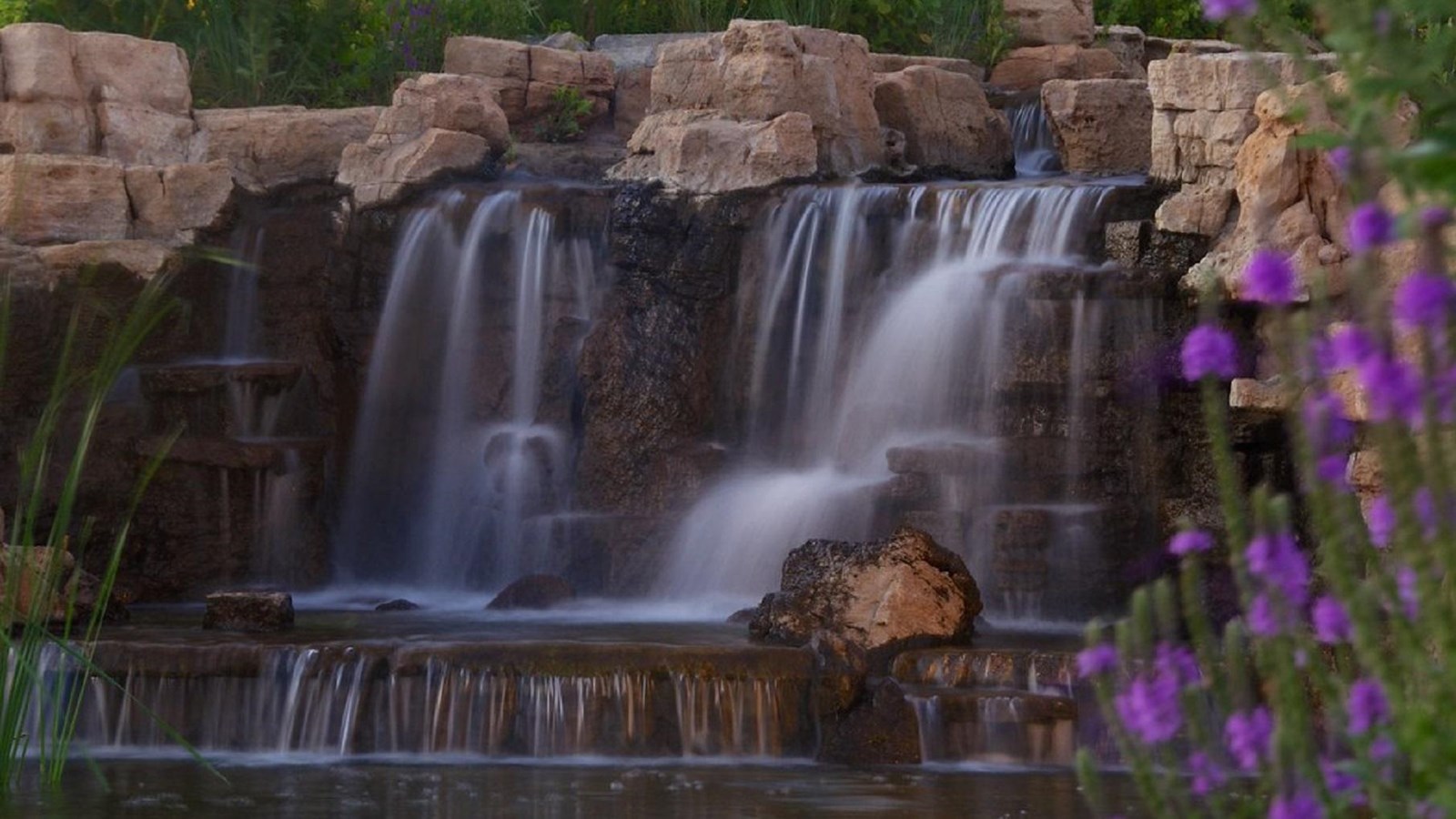 Water tumbles over sandstone to a pool below, surrounded by lush greenery and purple flowers