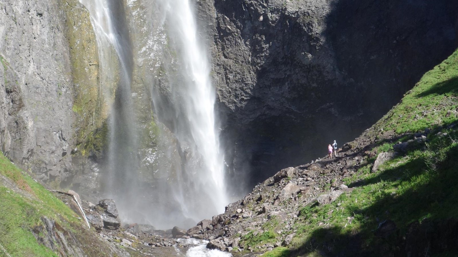 A white plume of water descends into a rocky canyon with hikers viewing from a trail. 