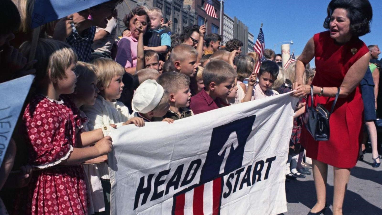 With school children, the first lady helps hold up a Head Start banner outside.