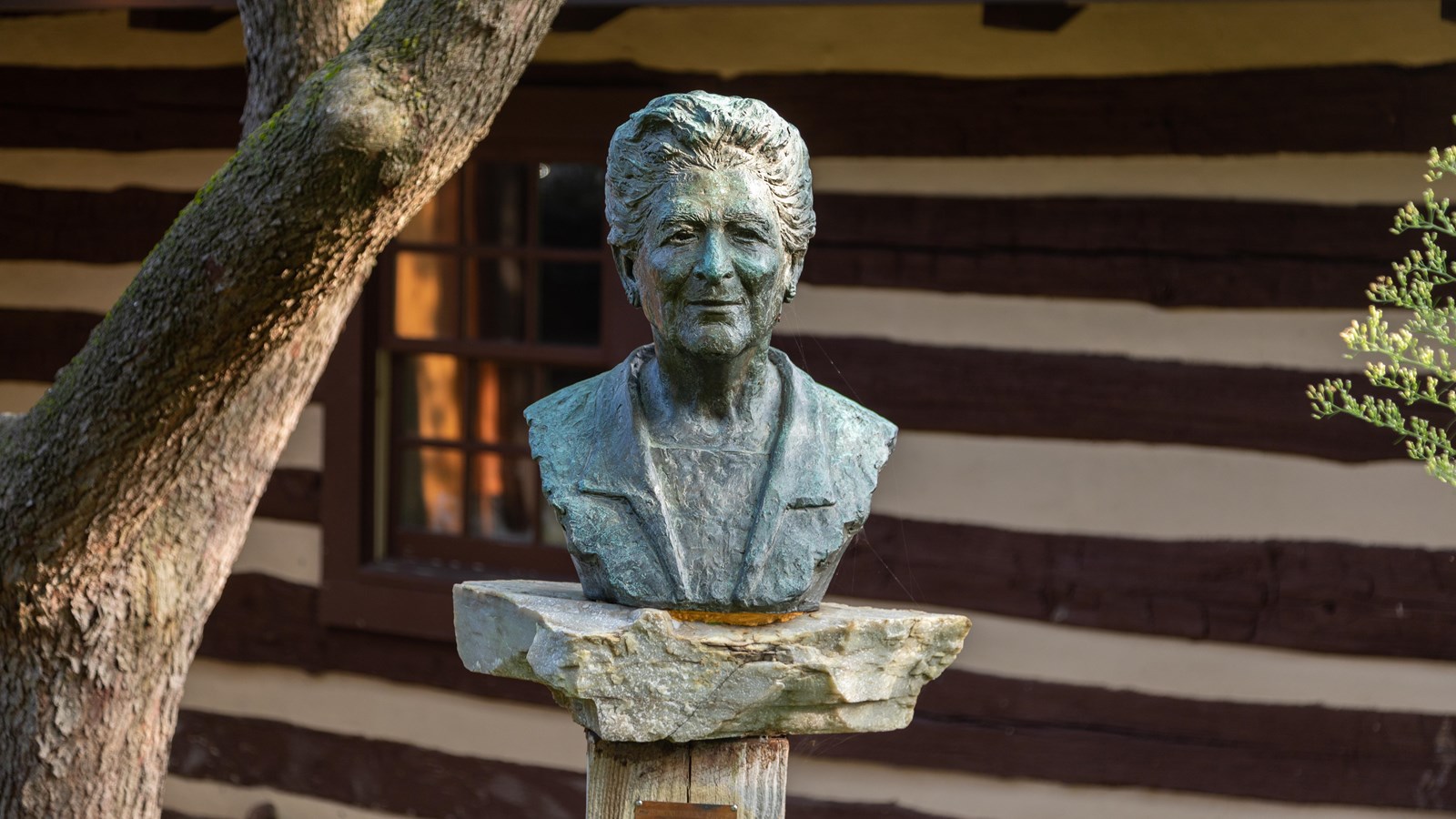 A bronze bust atop a stone slab on a wooden pedestal in front of a log cabin building.
