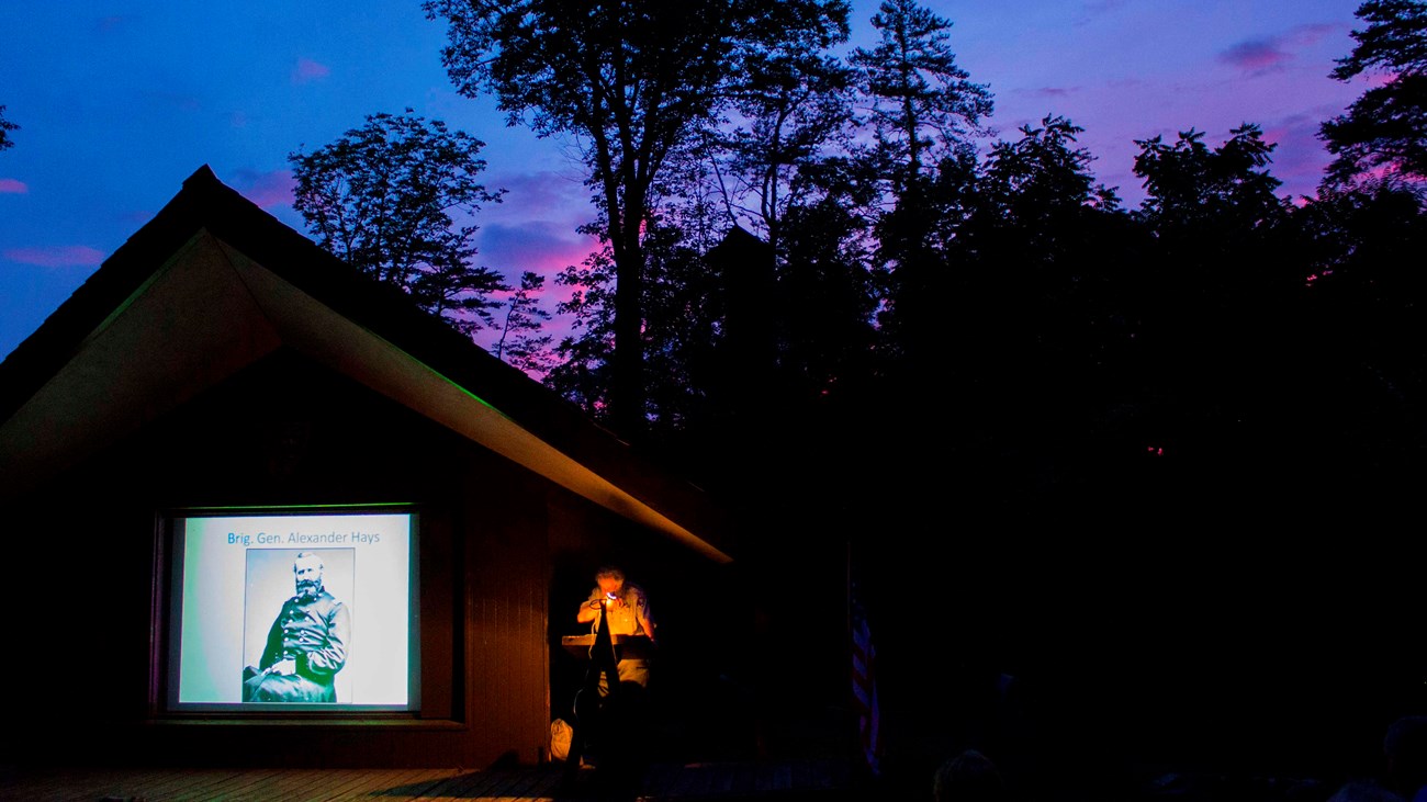 A park ranger at an amphitheater speaking with a slide show next to him.