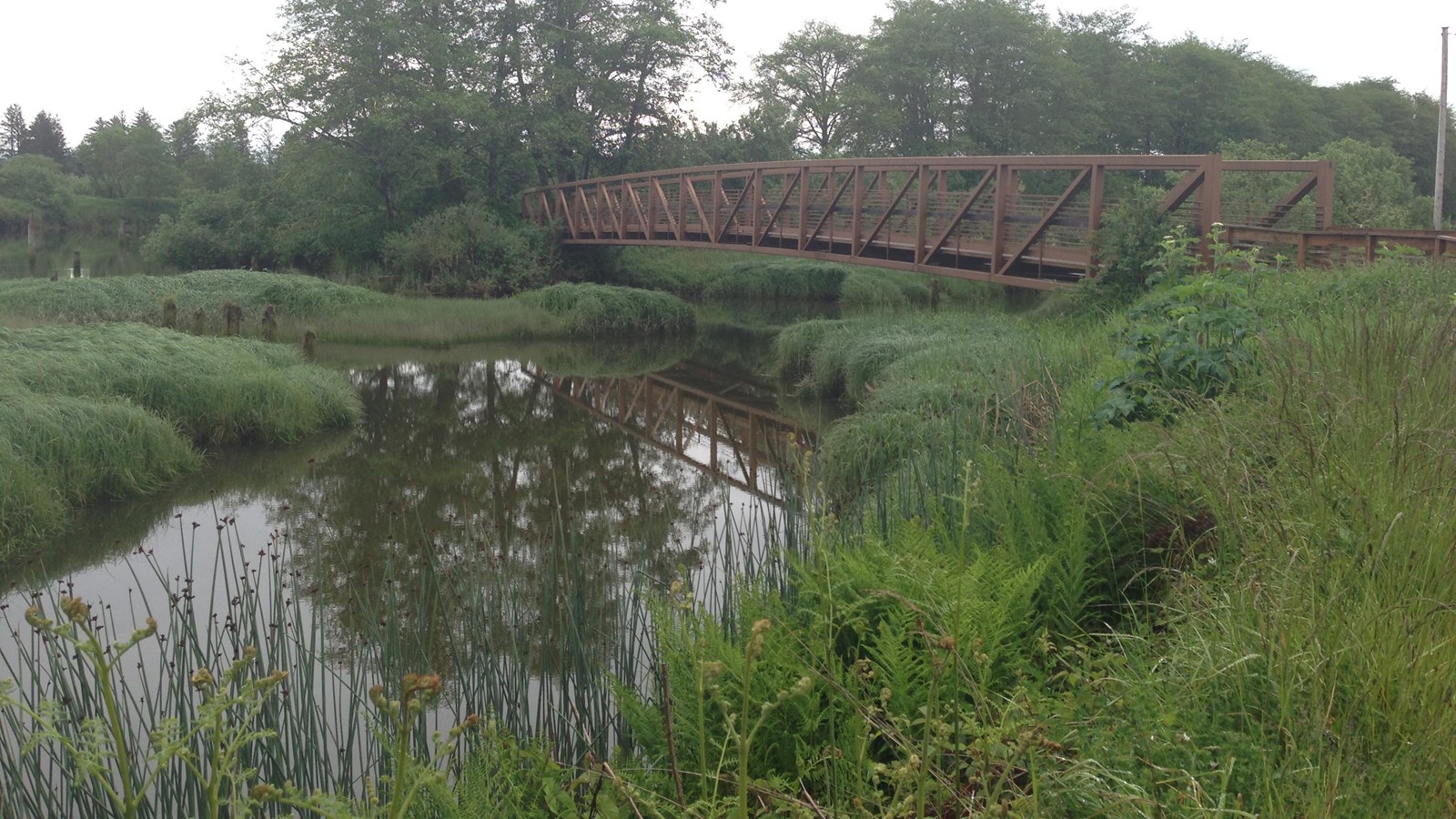 A brown metal bridge extends over a still river, the reflection of the bridge visible in the water
