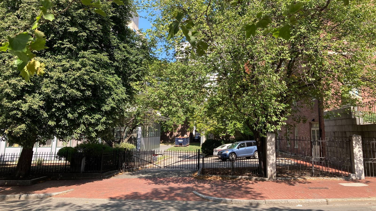 A neighborhood sidewalk with two trees on either side. A lot is behind the sidewalk.