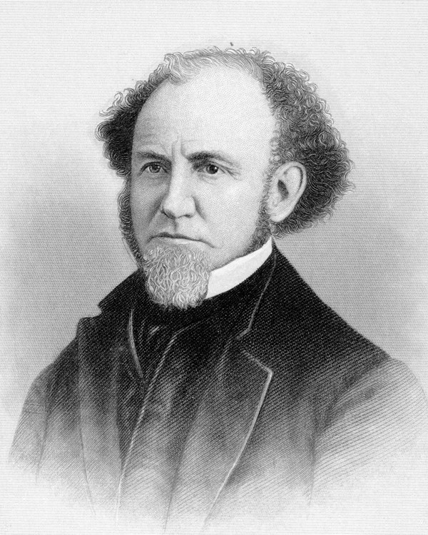 Bearded man with balding, curly hair whose white collar sticks up above his dark coat and vest.
