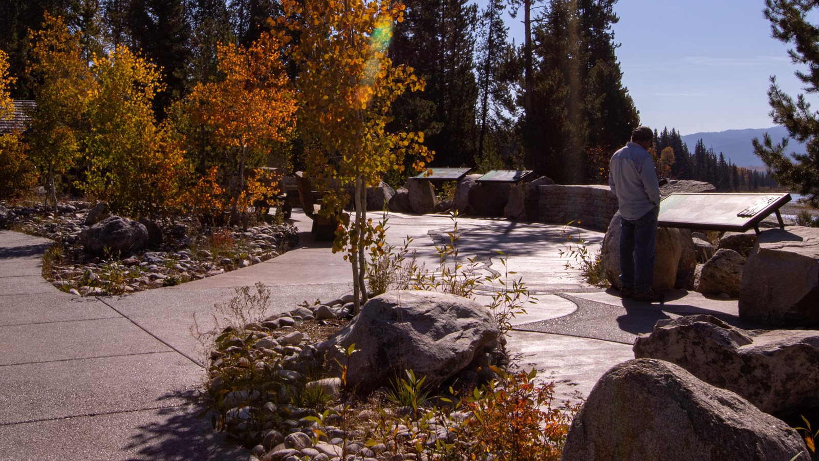 A man looks at a panel overlooking the river with a concrete plaza, trees, and rocks nearby. 