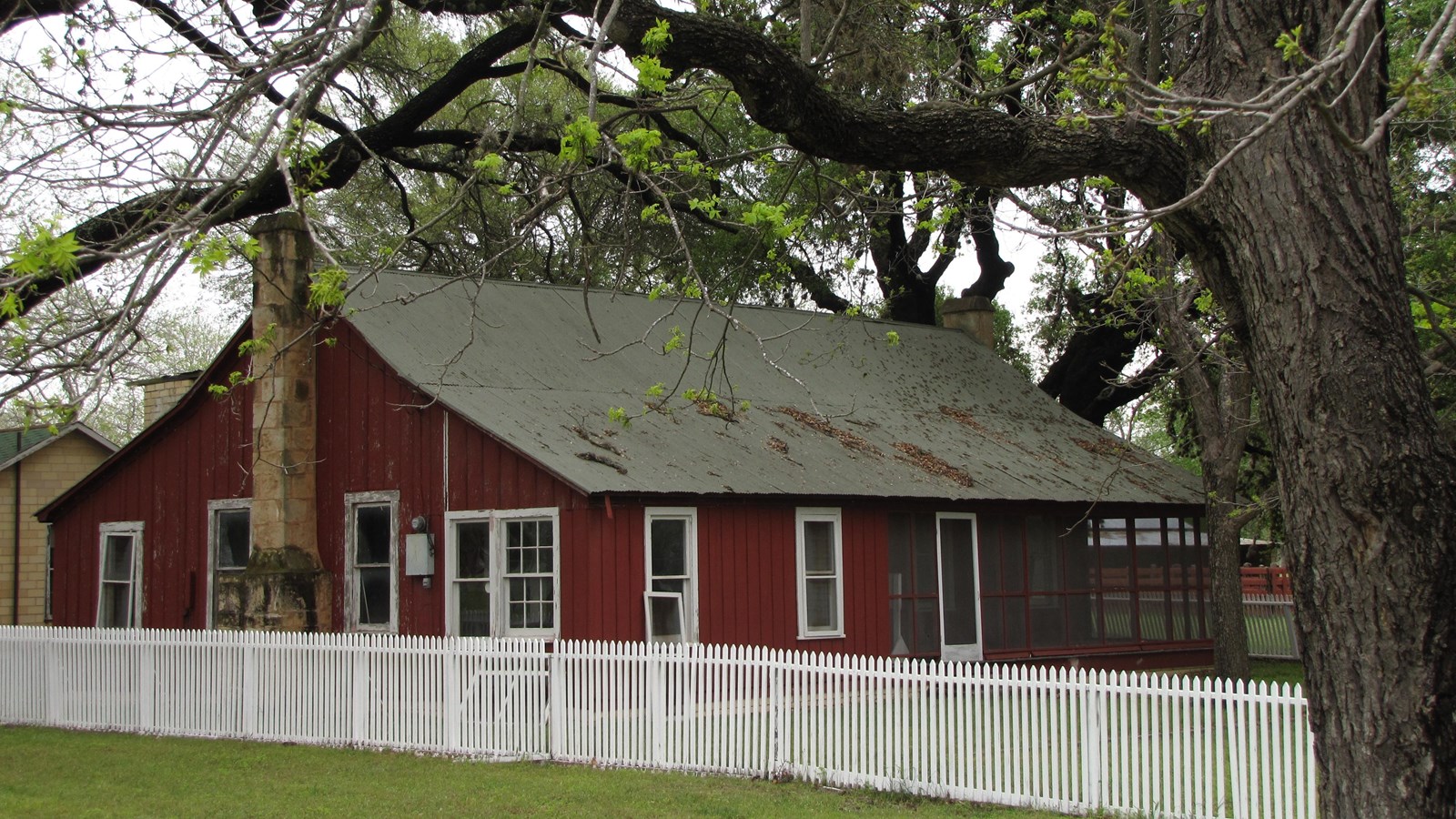 A red house with white-trim whites is behind a white picket fence, all under large shade trees.