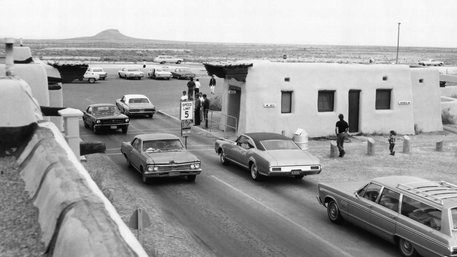 Vintage cars pass the adobe visitor center and bathrooms on a road with a butte in the background