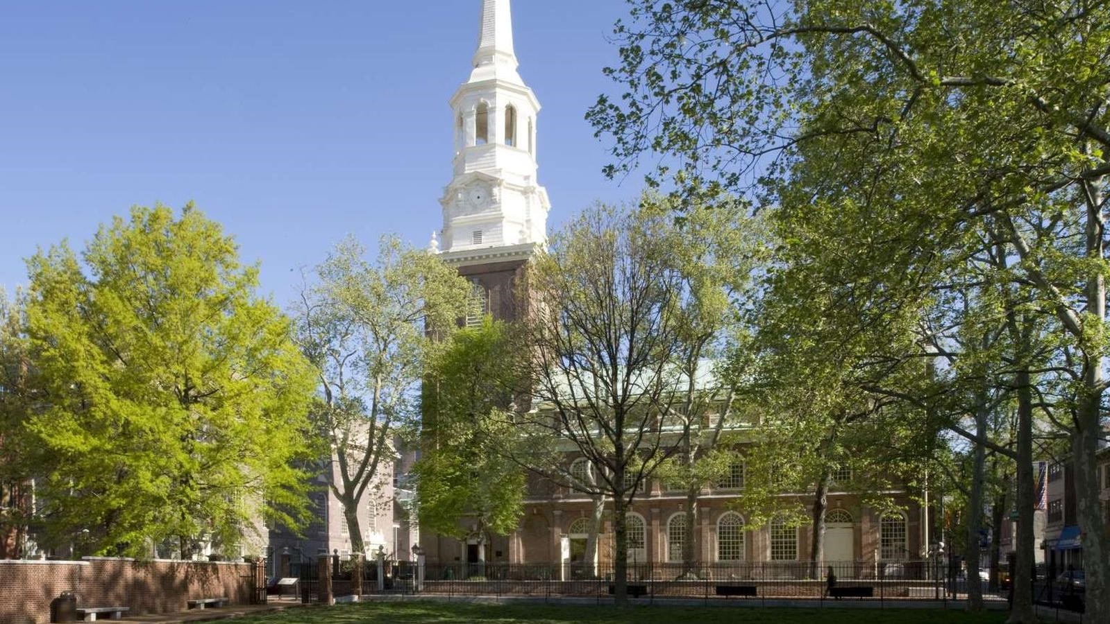 A color photo showing a red brick church with a tall white steeple with trees on the foreground.