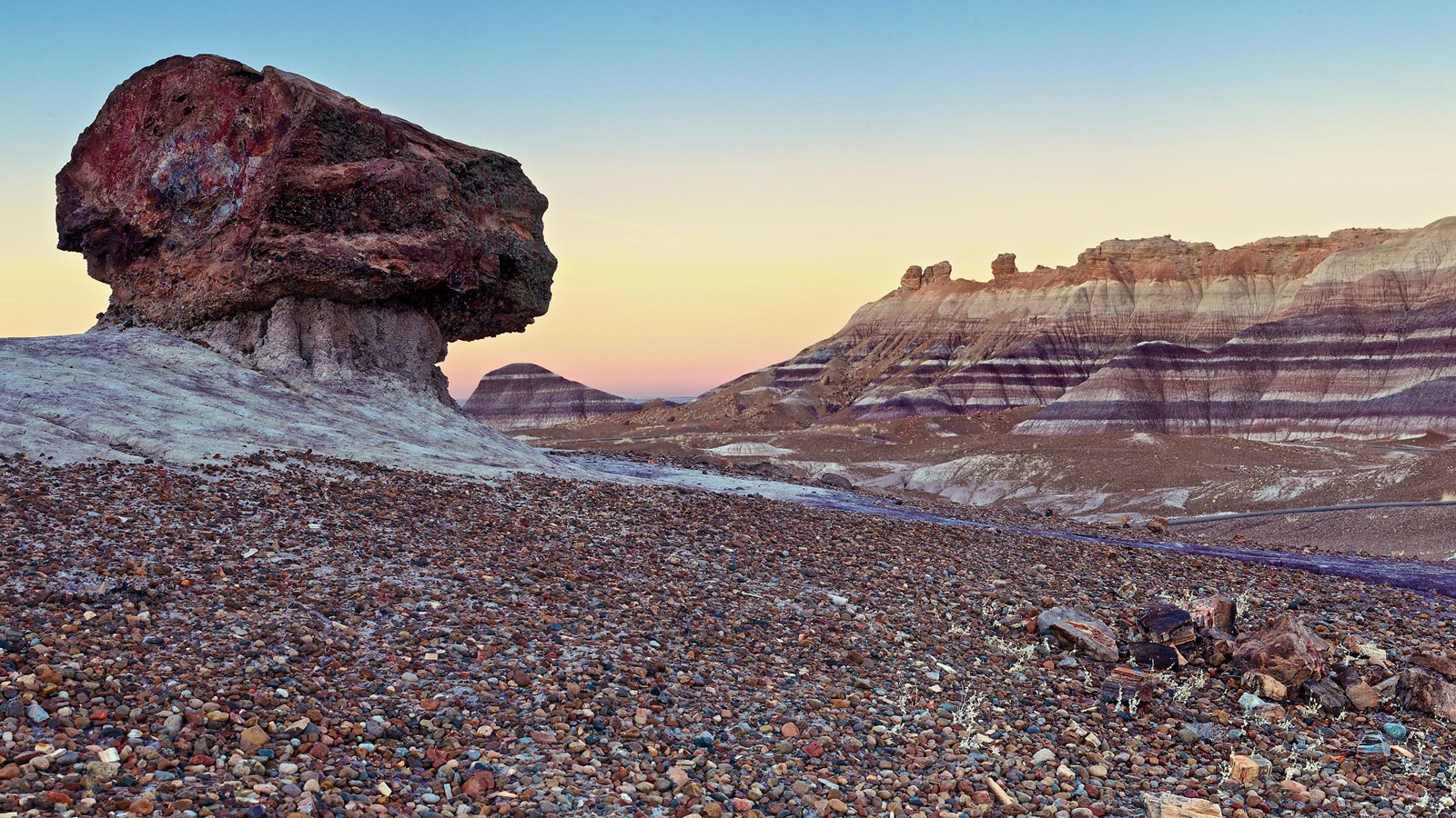 A pedestal log balances in front of a blue, purple, and gray banded badland at twilight.