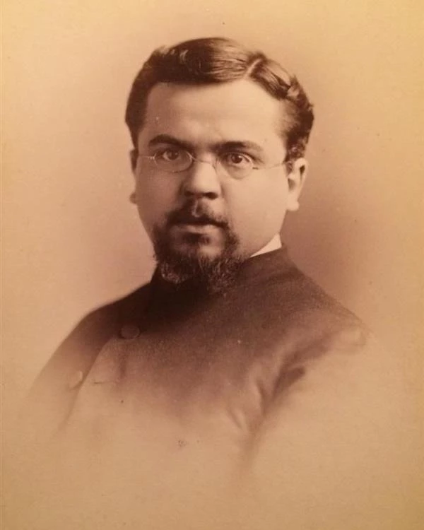 A man with dark hair, beard and glasses wears a dark jacket with a white shirt underneath.