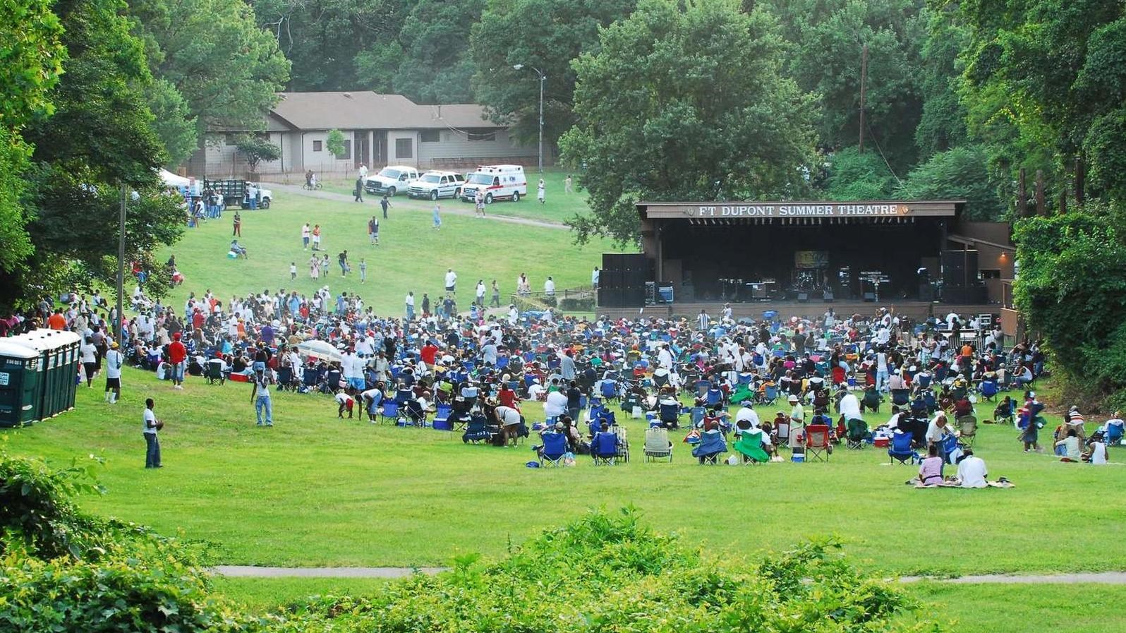 Several people are gathered around a stage and waiting for a summer concert to begin at Fort Dupont.