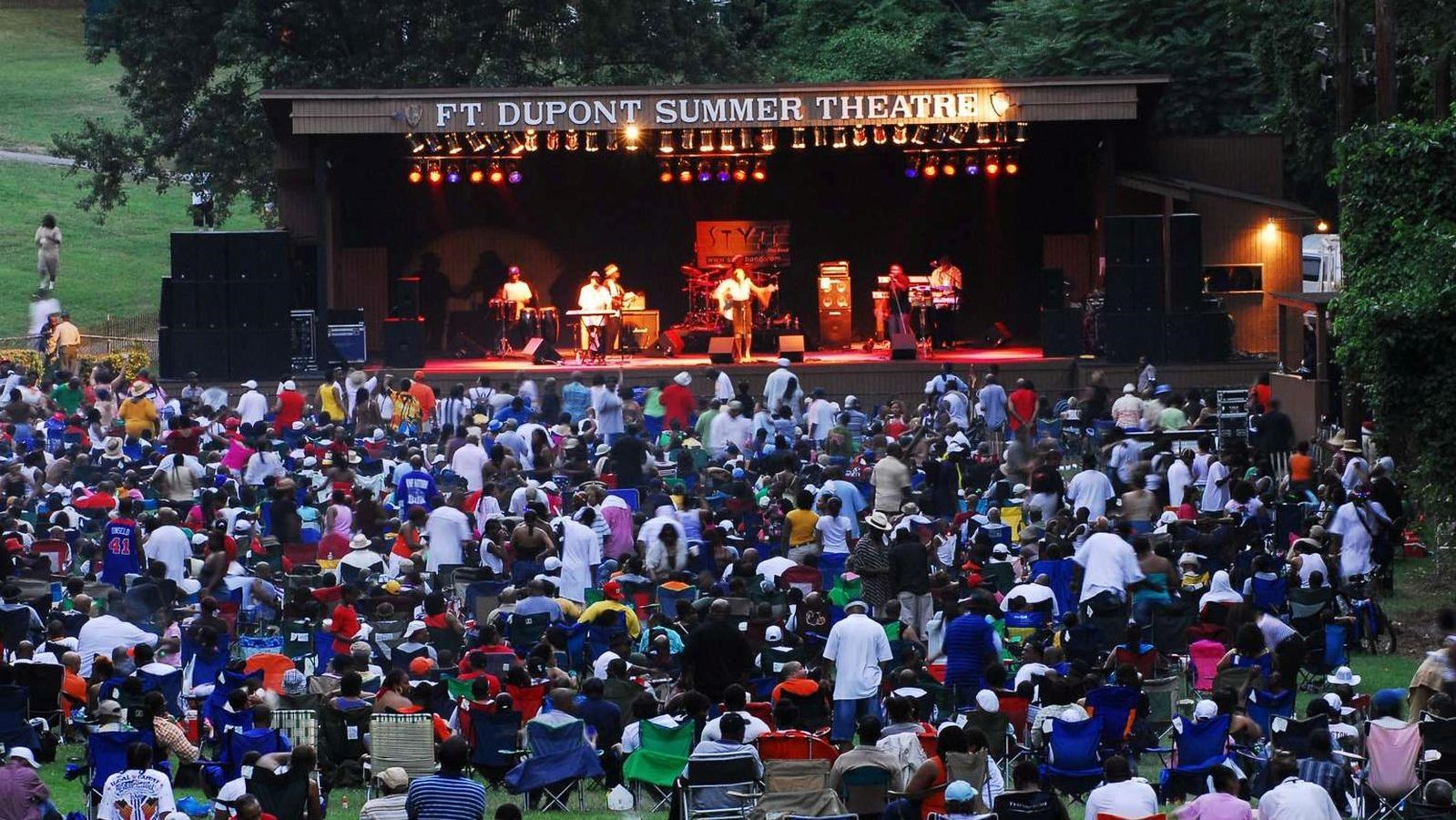 Several people are gathered around the Fort Dupont Summer Theatre stage for an evening summer concer