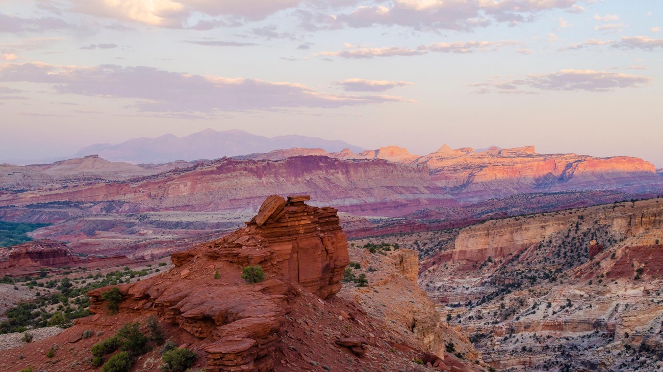 Colorful red rocks, striped cliffs, mountains in the distance, and a pale blue moving to purple sky.