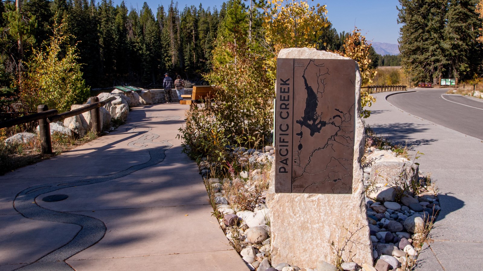 A plaza overlooking the river with a large rock with a bronze sign in the front with trees.