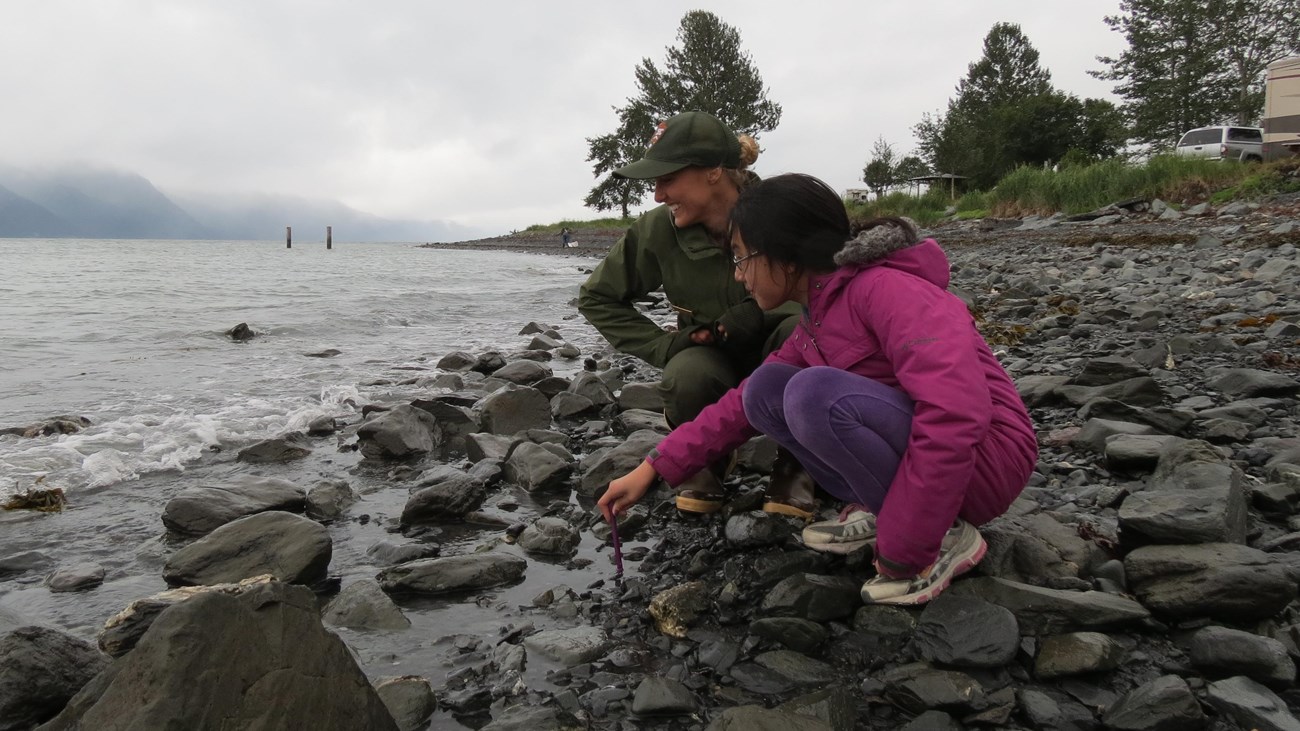 A park ranger and a child standing on a rocky beach. They are looking out into the water