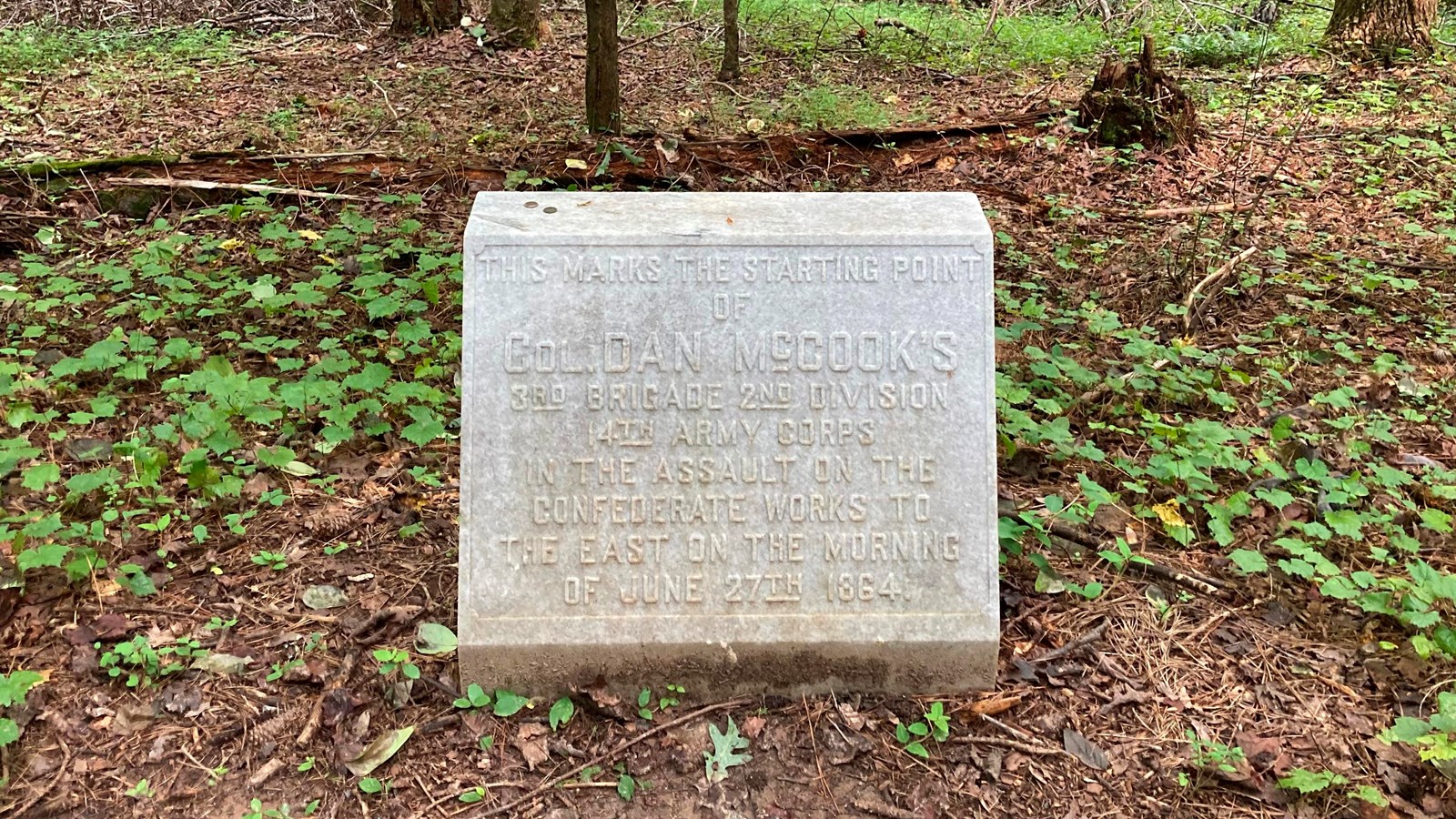 A square like marble marker sits in a small clearing to the right of a hiking trail. 