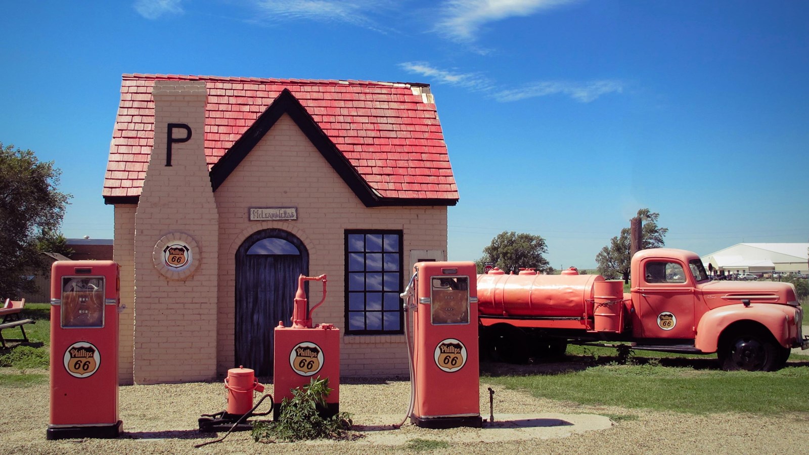 A small tan building with red roof. Out front are old red gas pumps and an old red pickup truck.