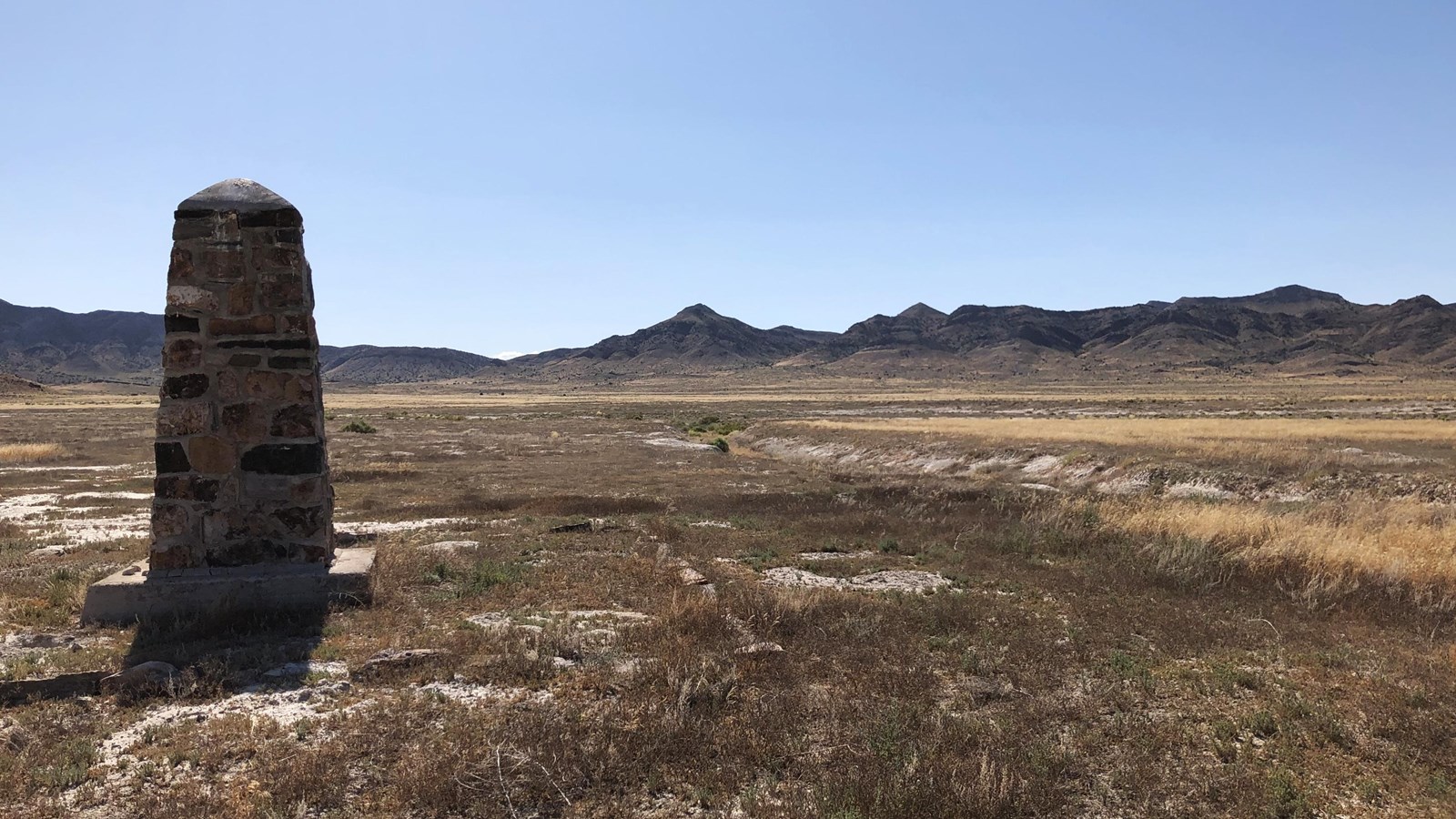 Stone obelisk sits by dry wash on barren plain with grass, distant mountains, and clear sky.