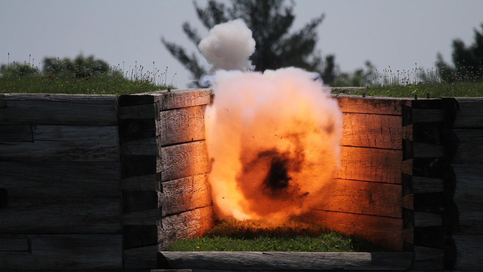 A ball of sire explodes from a cannon barrel out of a gap in the wooden fort wall. 