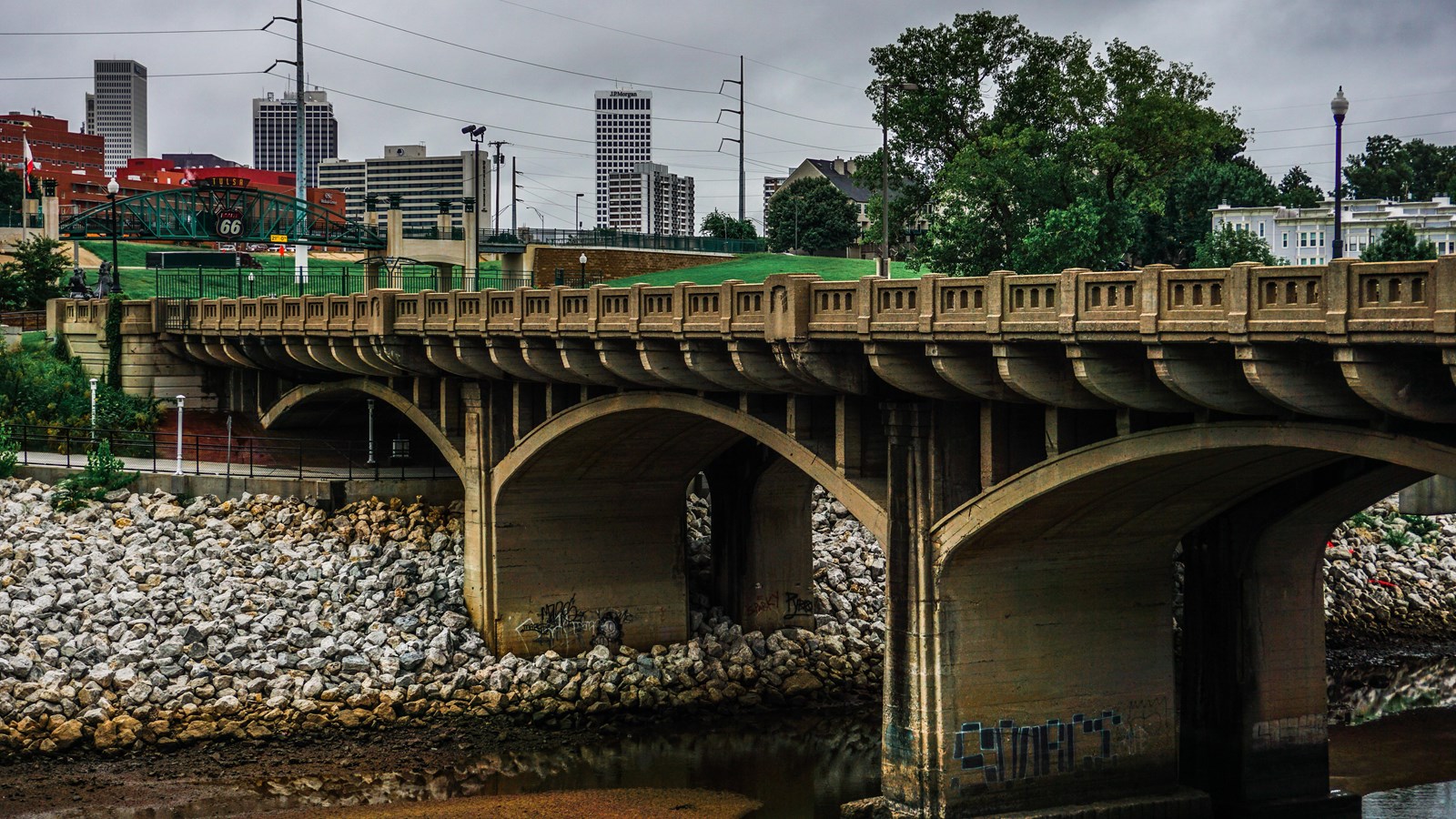 A large bridge with tan concrete supports. In the background are skyscraper buildings and trees.