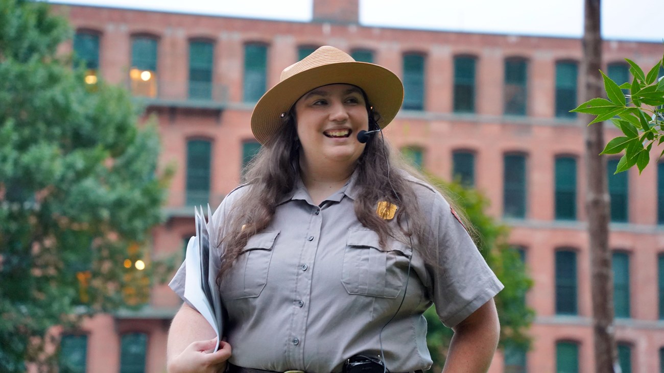 Ranger stands smiling holding papers in front of 4-story brick mill