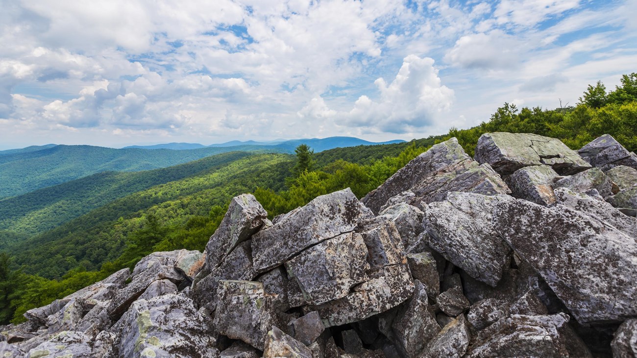 A jumble of large boulders sits on a cliff over a view of the valley below.