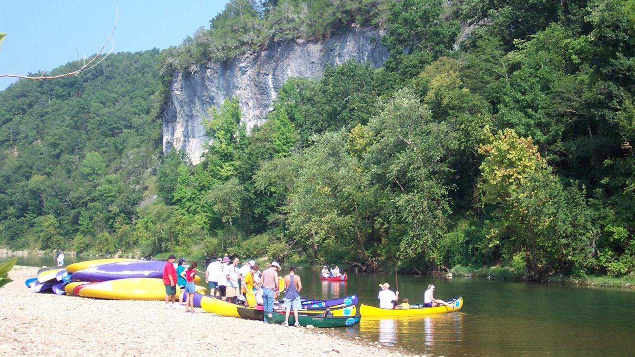 pile of canoes on gravel riverbank with trees and cliff in background