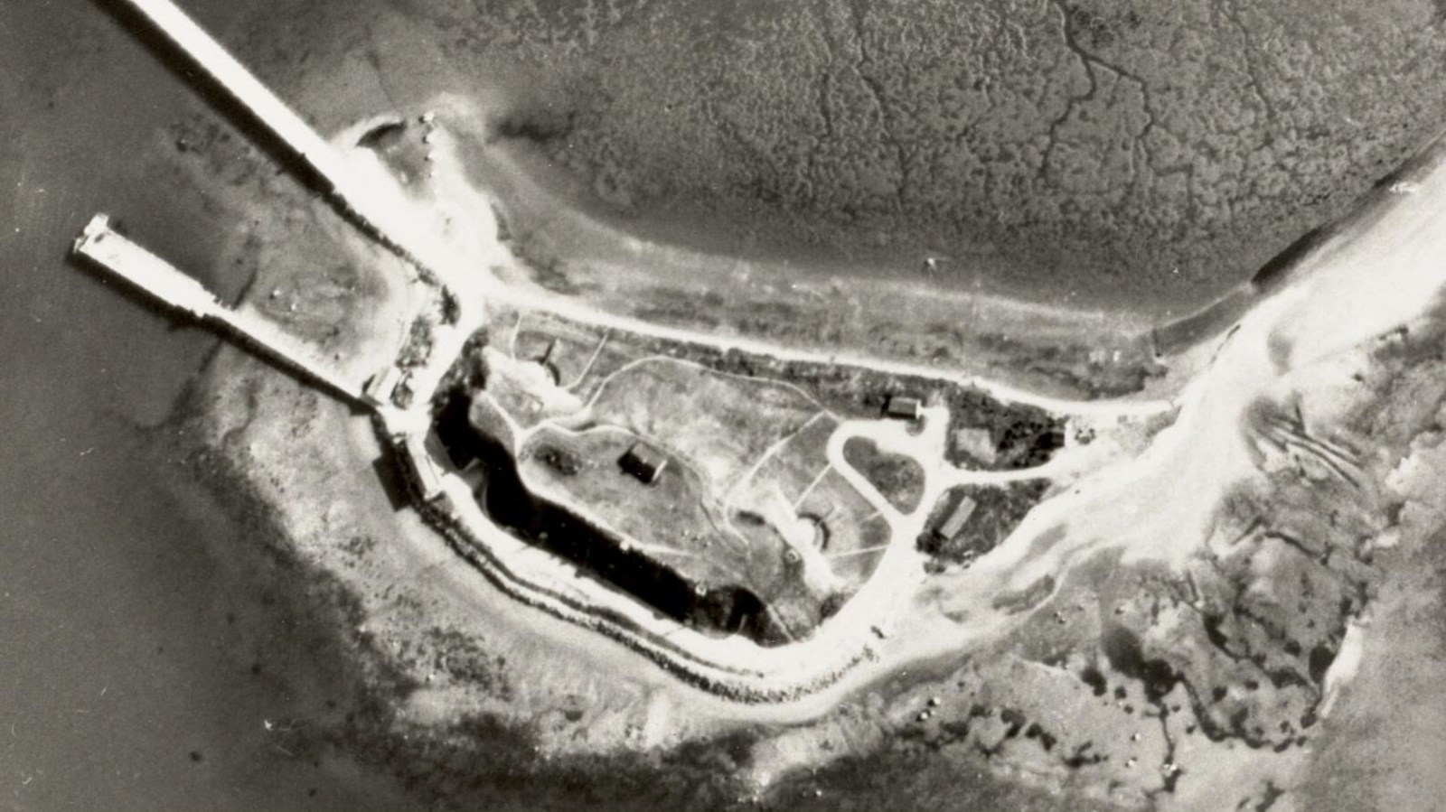 	A black and white aerial photo of an island. A narrow bridge stretches across the water.