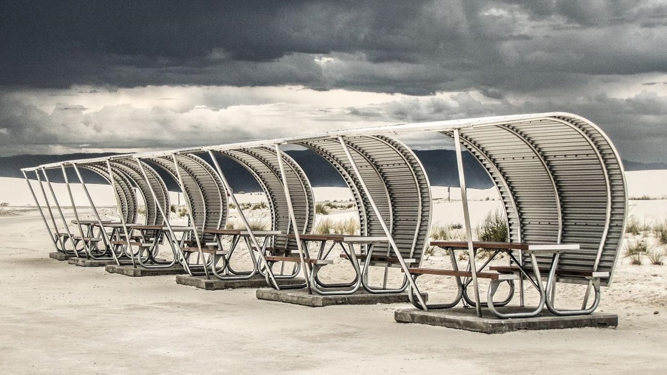 a row of curved metal structures provides shade to picnic tables on a concrete pad.