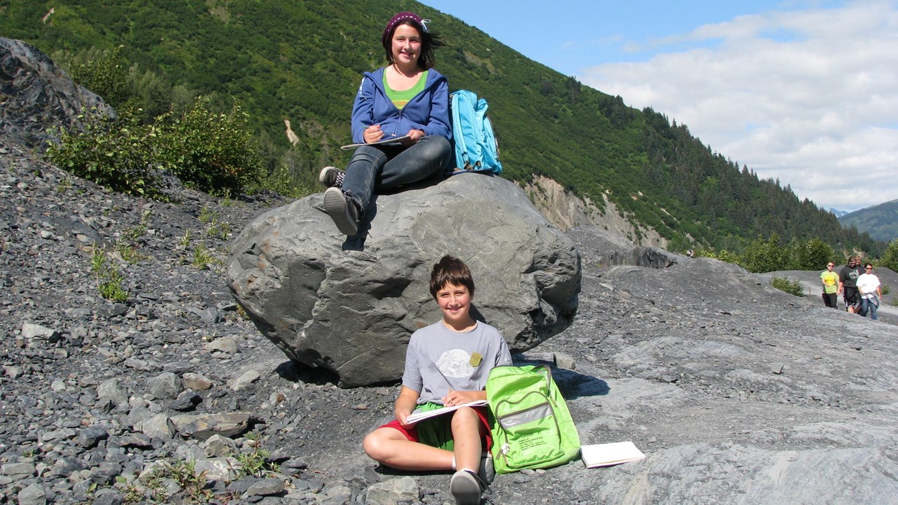 Two kids sitting down. One is on a boulder and the other on the ground.