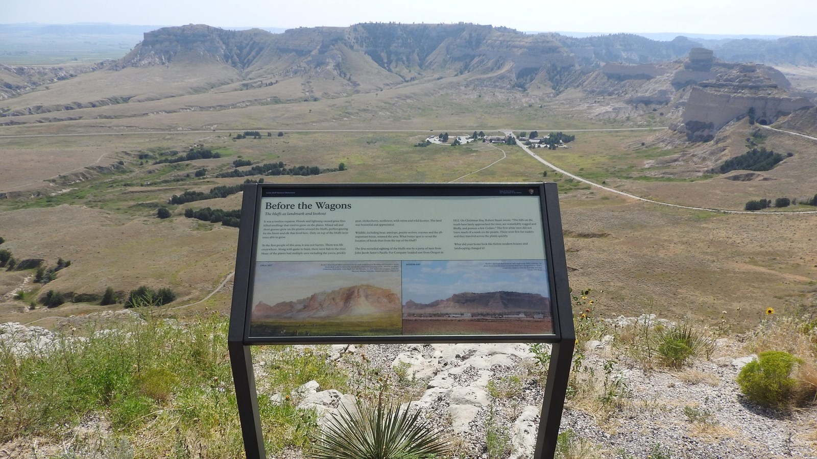 A wayside interpretive panel at an overlook of a sandstone bluff and a cluster of buildings.