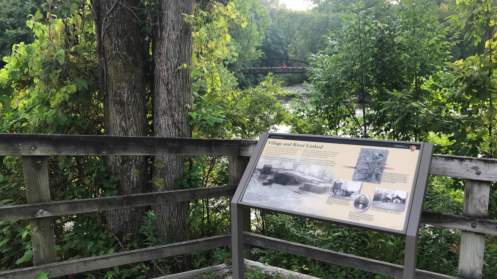 An exhibit by a wooden fence overlooking a trail bridge over a river.