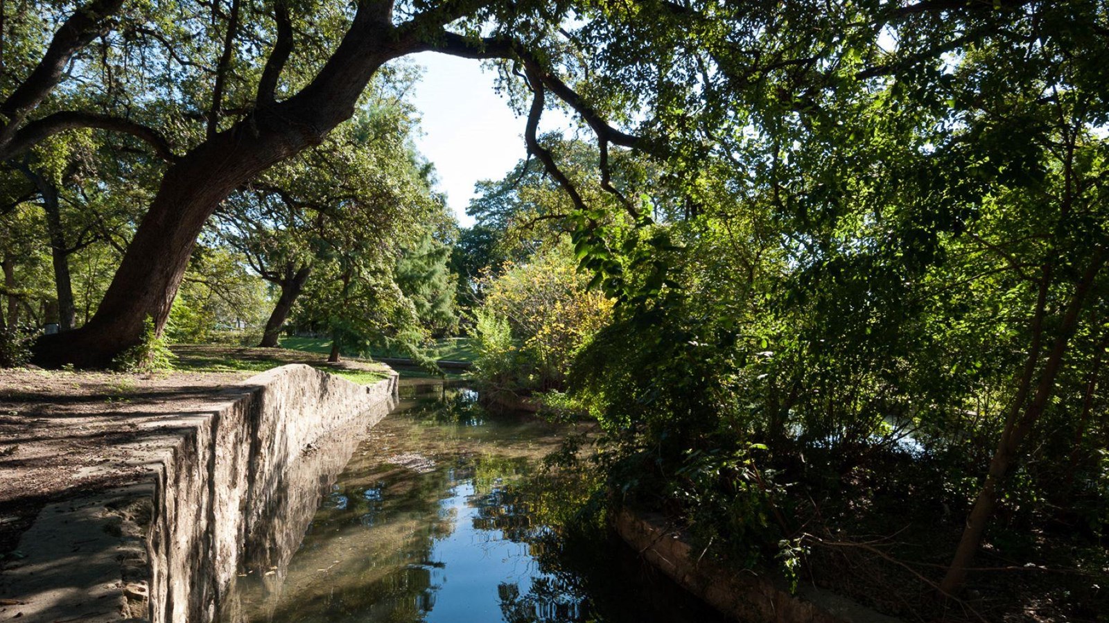 A stone wall runs next to a still irrigation ditch, with trees stretching over top.
