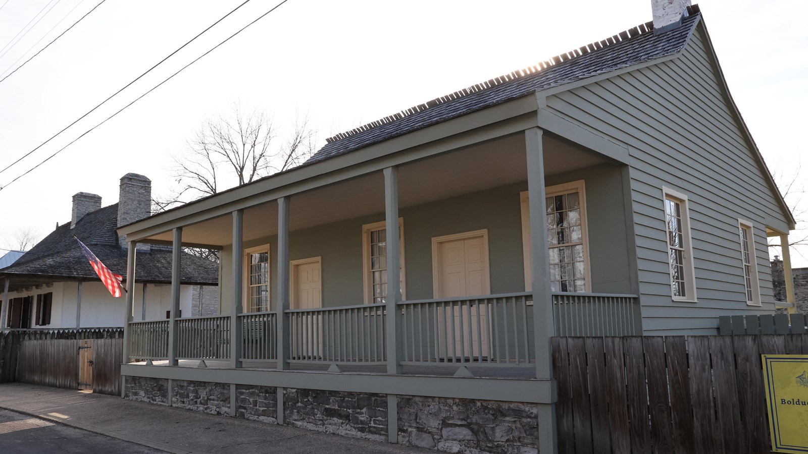 Green house with open front porch and American flag flying in the breeze, with a short brown fence.