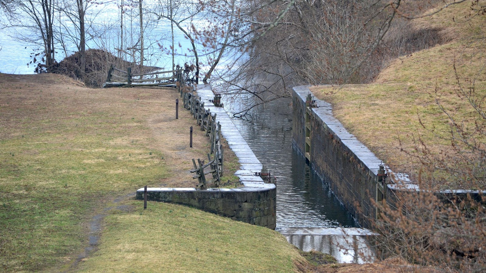 A narrow canal, bounded by stone walls, flows through a grassy field and into a river.
