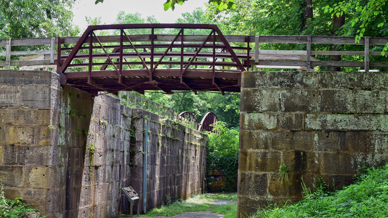Two brown metal trail bridges cross over a path that leads inside a stone canal lock to exhibits.
