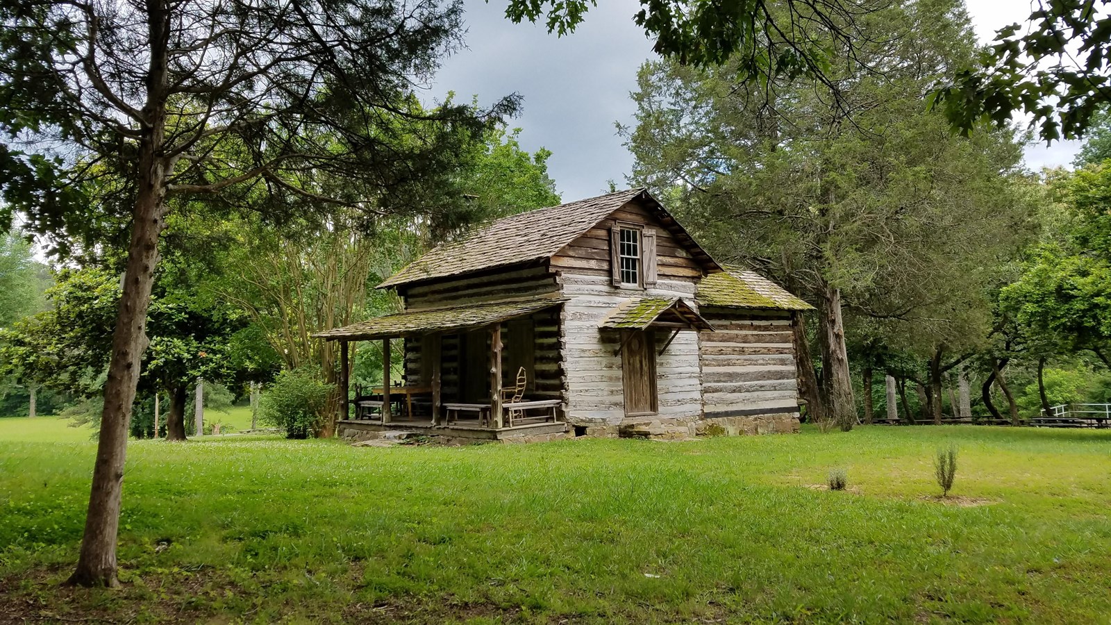 Historic log cabin sits in a field of grass surrounded by trees.