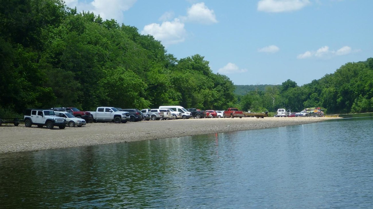 blue river in foreground with many cars parked on tan gravel at center