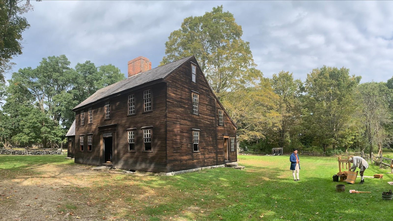 A salt box style house made of wood. People in 18th century clothing stand out front pressing cider