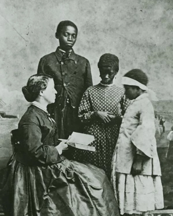 Laura sits with a book while three students stand in front of her.